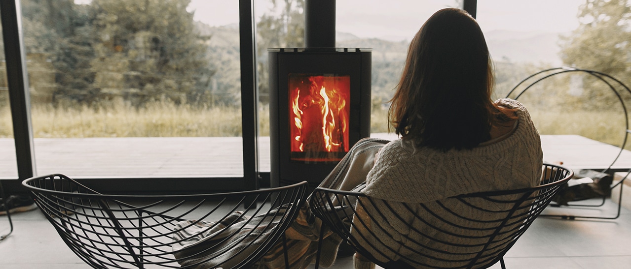 Frau genießt Wärme vor Kamin und blickt aus großen Fenstern in die Natur. 5 einfache Tipps zum Energie- und Heizkostensparen.