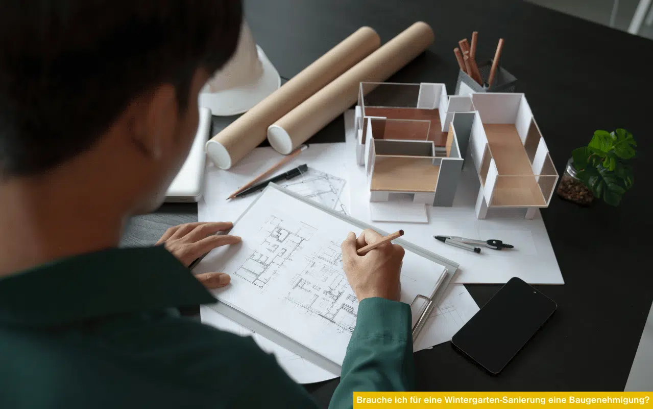 Brauche ich für eine Wintergarten-Sanierung eine Baugenehmigung zeigt Person beim Zeichnen von Bauplänen und Modell auf dem Tisch
