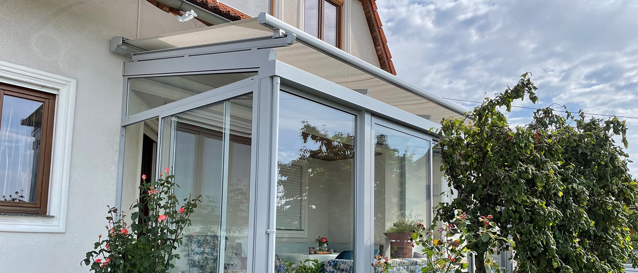 Das Bild zeigt einen Wintergarten an einem Wohnhaus mit Glasfronten, umgeben von Pflanzen und Rosen, bei sommerlichem Wetter.