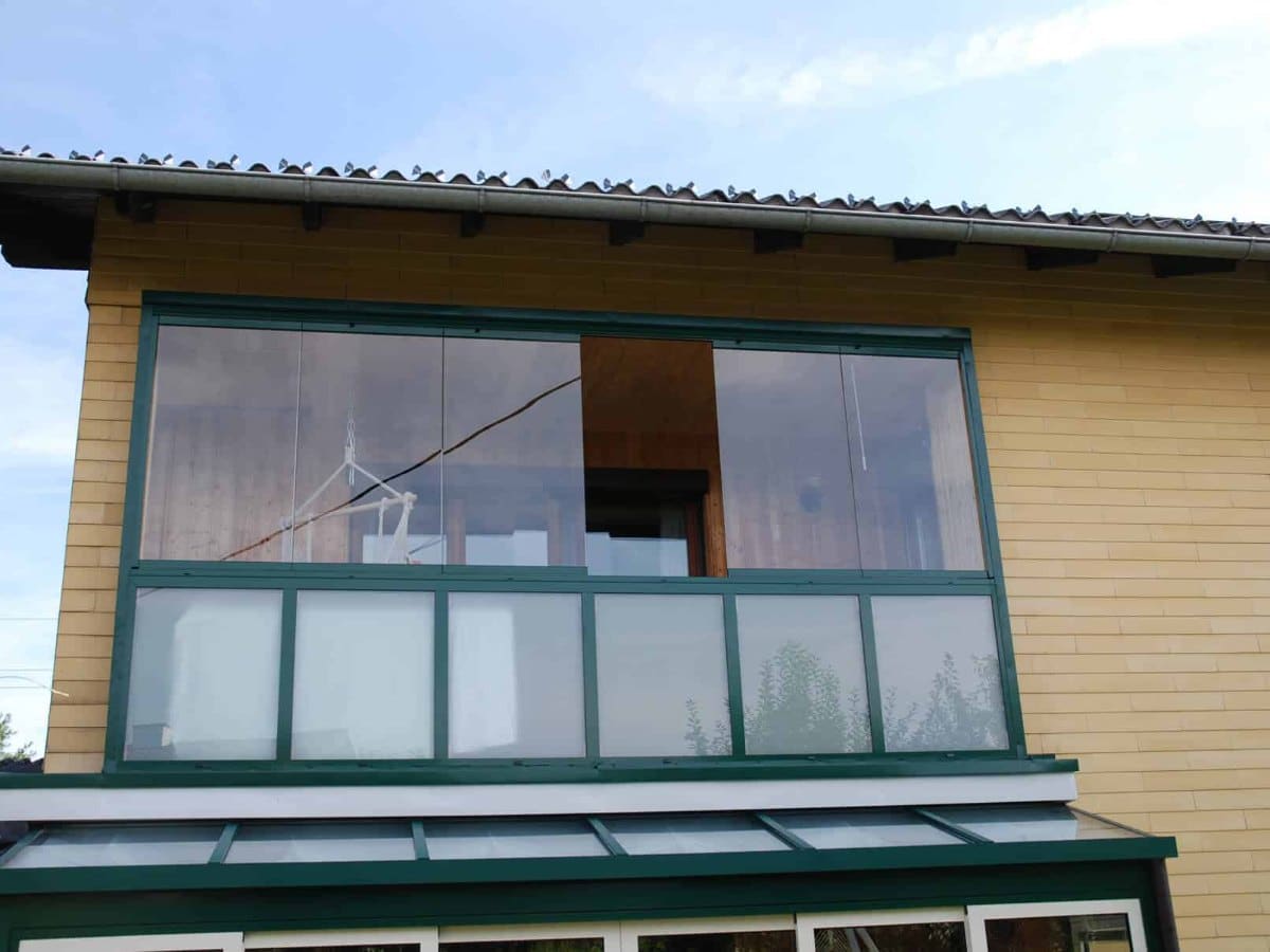 Balkonverglasung mit rahmenlosen Faltfenstern und grünen Rahmen an einem Holzhaus