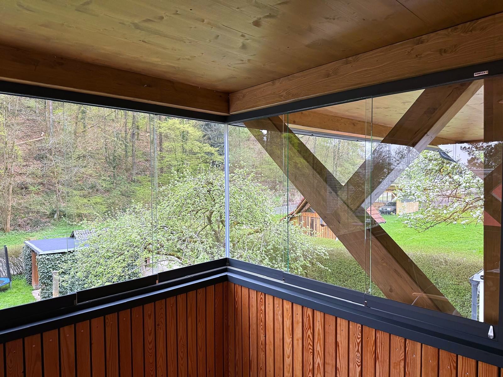 Balkonverglasung mit rahmenlosen Ganzglas-Schiebefenstern auf einer Brüstung aus Lärchenholz unter Holzdecke, Eckverglasung, Oberösterreich