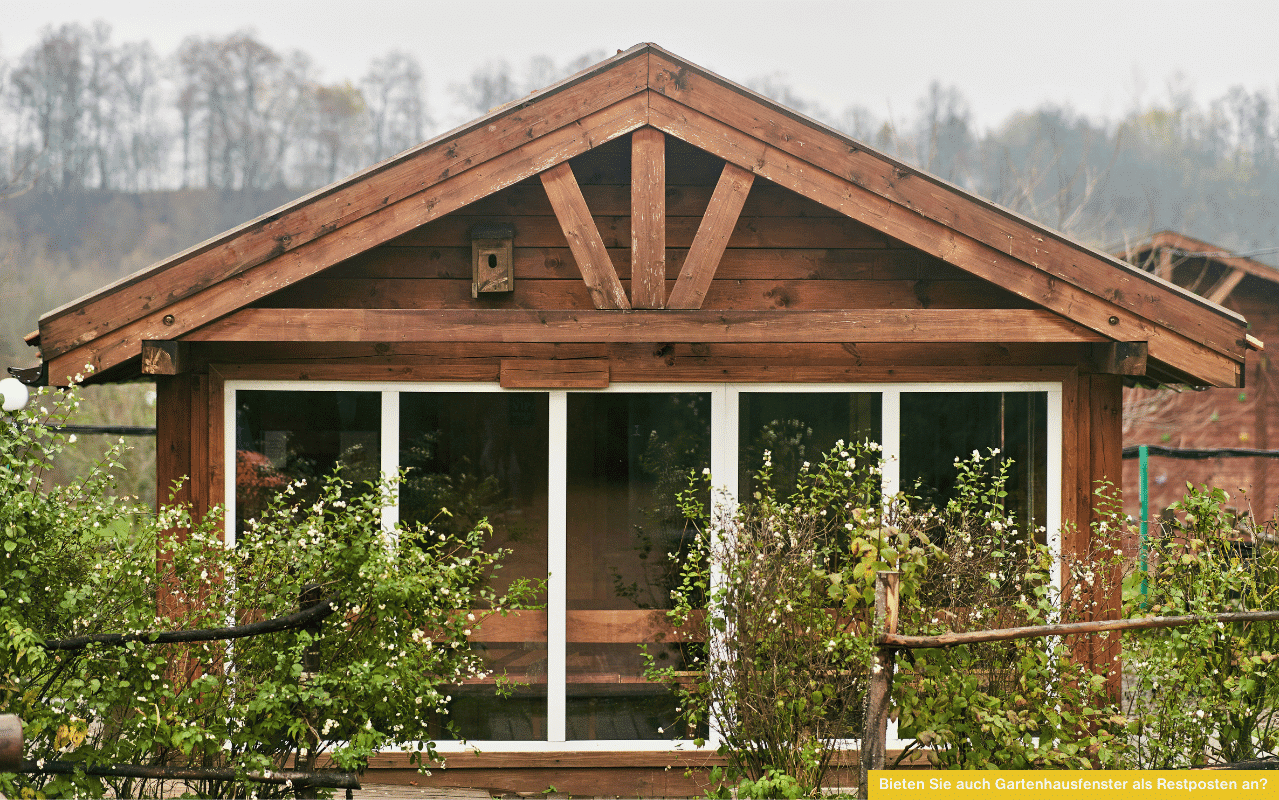 Bieten Sie auch Gartenhausfenster als Restposten an? Blick auf ein kleines Gartenhaus aus Holz mit großem Fenster und Blumen davor.