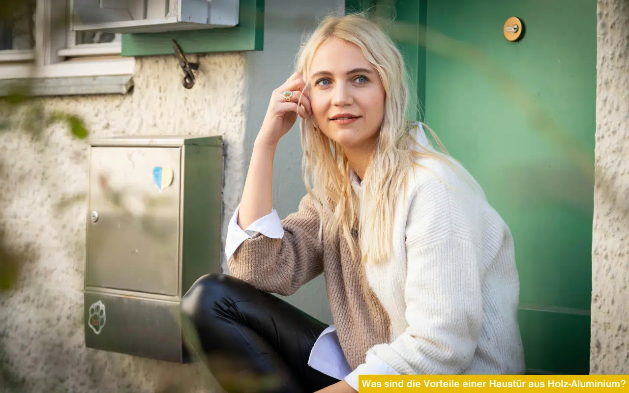 Blonde Frau sitzt vor grüner Haustür. Was sind die Vorteile einer Haustür aus Holz-Aluminium? Wetterfester Sonnenschutz.
