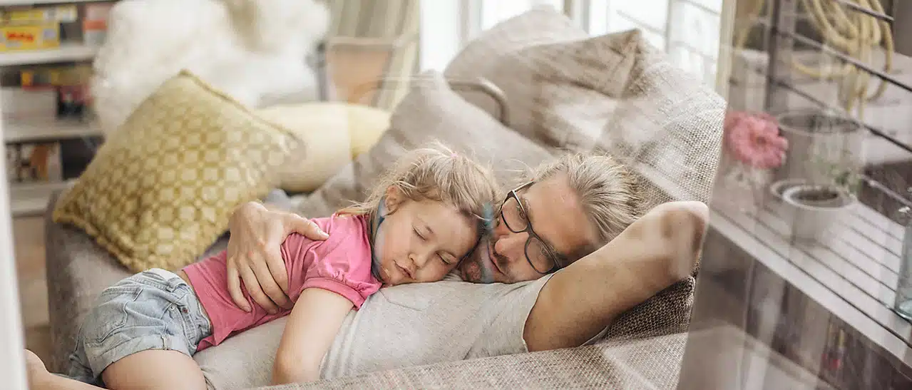 Ein junger Mann mit seiner Tochter schläft bequem auf dem Sofa. Sie sind dank Schallschutzfenstern bestens vor Lärm geschützt. Bringen Schallschutzfenster etwas - das klären wir in diesem Beitrag!