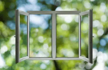 Geöffnetes Doppelflügelfenster mit klaren Linien, im Hintergrund unscharfe grüne Bäume und Sonnenreflexe, die für Frische und Weite stehen.