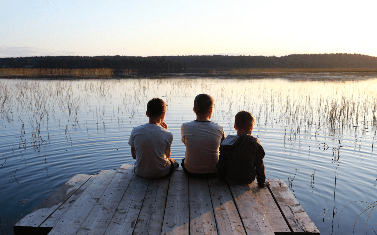 Drei Kinder sitzen bei Sonnenuntergang auf einem Steg am See – Symbolbild für gemeinsame Auszeit am Zwickeltag am Freitag, 20.06.2025.