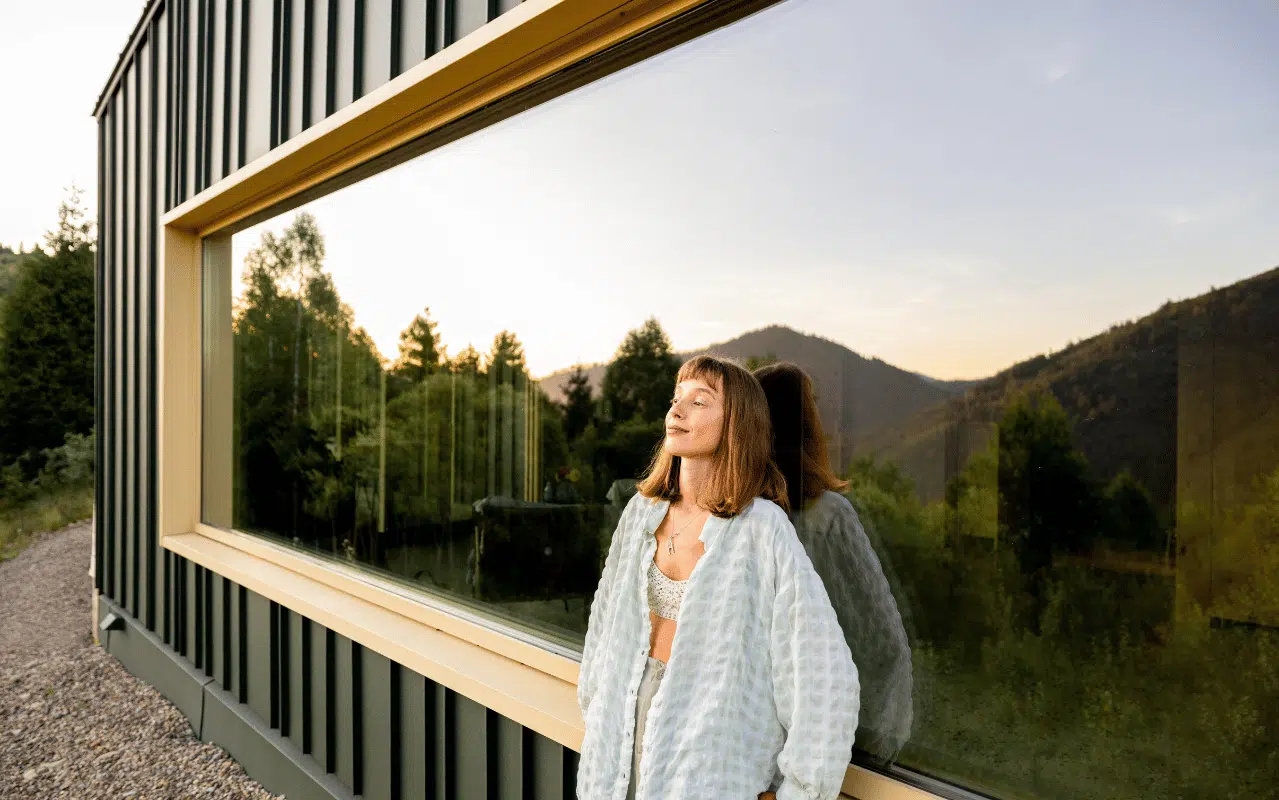 Vertrauen Sie auf Fenster Hersteller in Österreich - Eine Frau lehnt sich entspannt an ein großes modernes Fenster eines Hauses in den österreichischen Bergen bei Sonnenuntergang.