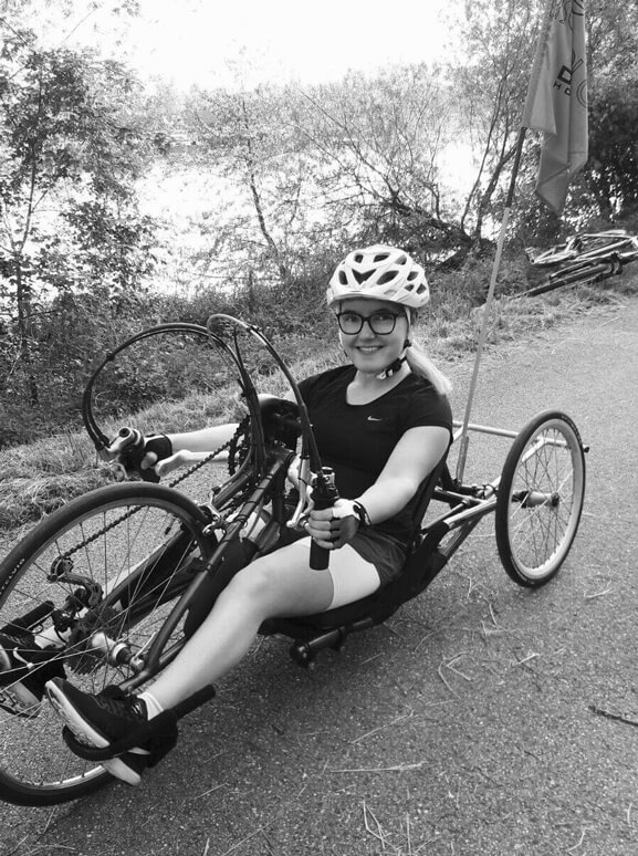 Das Bild zeigt Elisabeth Egger auf einem Handbike, sie trägt Fahrradhelm und Brille, sitzt entspannt auf einem Weg im Grünen und lächelt in die Kamera. Die Stimmung wirkt sportlich und positiv.