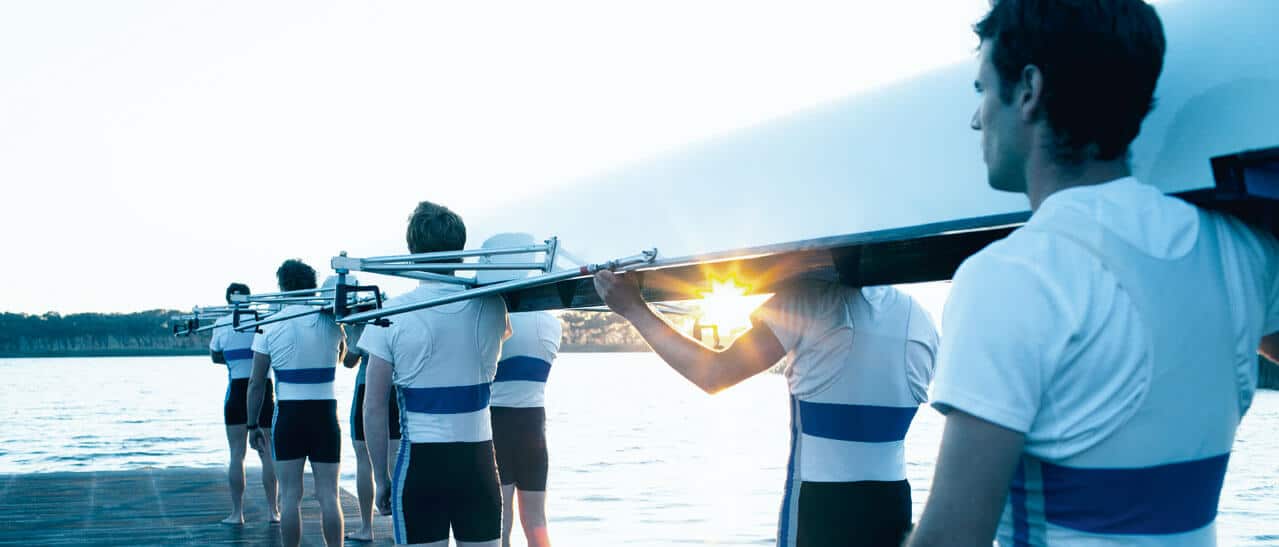 Exzellenz im Management erfordert Teamgeist und Präzision. Die Ruderer, die ihr Boot gemeinsam zum Wasser tragen, symbolisieren den kollektiven Einsatz, der notwendig ist, um in Unternehmen nachhaltigen Erfolg und Höchstleistungen zu erzielen.