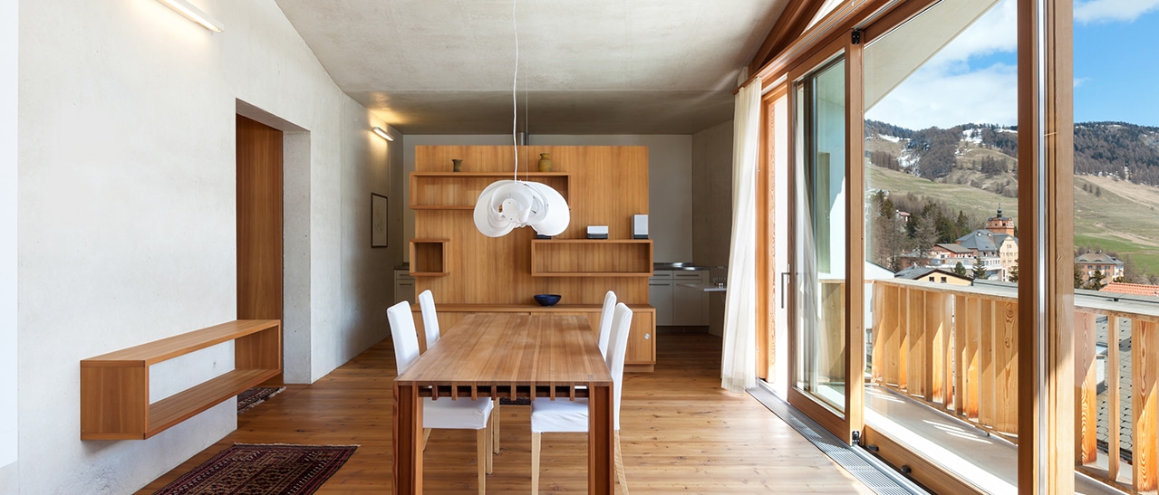Man sieht ein helles Esszimmer mit Holzboden, einem Tisch mit Stühlen und großen Holzfenstern bzw. einer Holz-Schiebetür zum Balkon. Draußen ist Berg- und Ortsblick in Österreich.
