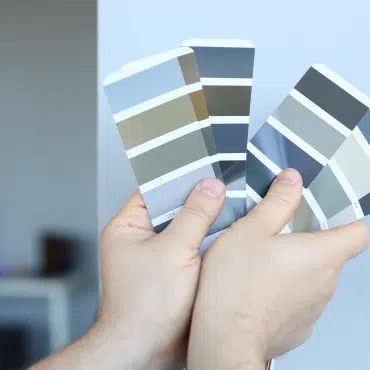 Fenster in RAL 7023 Betongrau, zwei Hände halten eine RAL-Farbkarte mit Grautönen und passender Farbe.