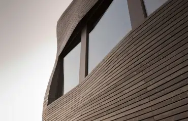 Eine Fassade aus Holz mit Fenstern in RAL 7013 Braungrau zeigt, wie sich moderne Farbwahl und natürliche Materialien perfekt ergänzen. Der warme Braun-Grauton verleiht dem Gebäude Tiefe und stilvolle Eleganz.