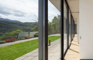 Das Bild zeigt eine moderne Glasfront mit bodentiefen Fenstern in RAL 7015 Schiefergrau. Die dunklen Aluminiumrahmen kontrastieren elegant mit der hellen Fassade und öffnen den Raum zur sonnigen Terrasse und Naturkulisse.