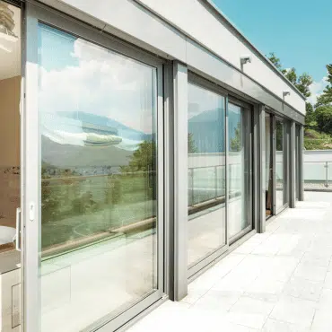 Das Bild zeigt eine moderne Glasfront mit mehreren großen Fensterelementen in RAL 7035 Lichtgrau. Die Aluminiumrahmen reflektieren das Sonnenlicht, während der Blick nach außen auf Berge und Landschaft fällt.