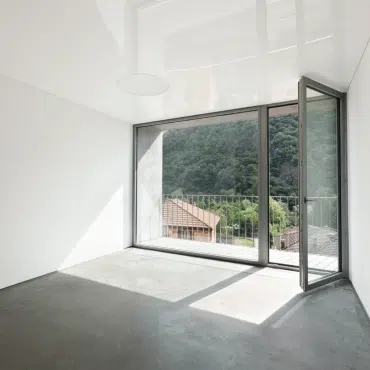 Das Bild zeigt ein großes, bodentiefes Fenster in der Farbe RAL 7038 Achatgrau. Der Rahmen ist modern und schlicht, das Fenster öffnet sich auf einen Balkon mit Blick auf bewaldete Hügel und Dächer.