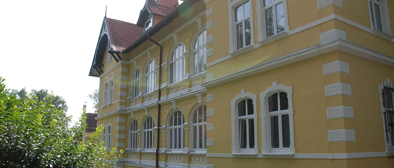 Fenster mit waagrechtem Kämpfer und Oberlichte in gelber Altbaufassade mit weißem Stuckrahmen und dekorativen Rundbogenformen.