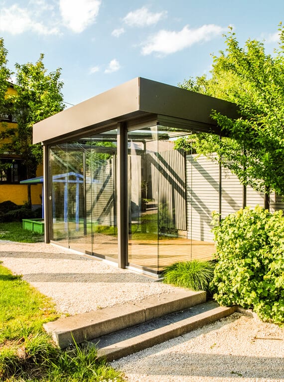 Blick auf einen modernen Glasanbau, der als eigener Baukörper im Garten steht. Große Glasflächen mit dunklen Rahmen umschließen einen Raum mit Holzboden, das flache Dach ragt seitlich hervor. Stufen und ein Kiesweg führen hinauf, rundum wachsen Sträucher und Bäume unter blauem Himmel.