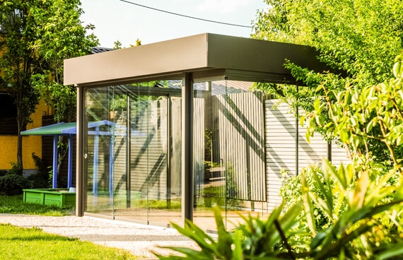 Ein freistehender Sommergarten im Garten, mit auskragendem Flachdach und schlanken anthrazitfarbenen Profilen. Die Seiten bestehen aus rahmenlosen Ganzglas-Schiebeelementen, in denen sich Sonne, Bäume und eine helle Holzwand spiegeln. Ringsum Rasen und Sträucher.
