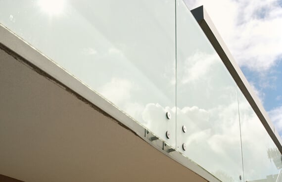 Nahaufnahme eines Windschutzes aus satiniertem Glas an einer Terrasse oder einem Balkon. Die großflächigen Glasscheiben sind mit runden Punkthaltern an der Konstruktion befestigt und bieten Schutz vor Wind und Einblicken.