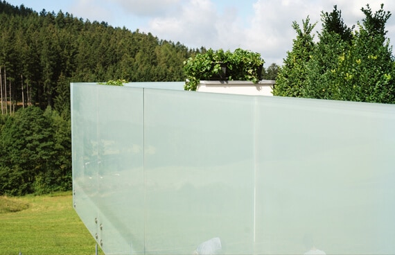 Großflächiger Wind- und Sichtschutz aus mehreren satinierten Glasscheiben entlang einer Terrasse. Die milchigen Glasflächen schränken Einblicke deutlich ein. Im Hintergrund sind grüne Wiesen, Bäume und ein bewaldeter Hügel zu sehen.