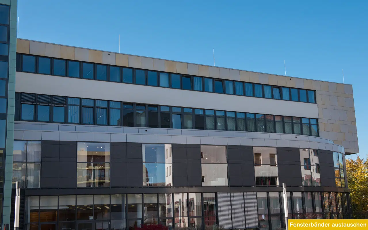 Große Glasfassade eines Bürogebäudes mit durchlaufenden Fensterbändern und modernen dunklen Profilen unter blauem Himmel.