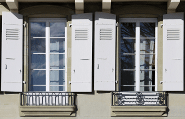 Zwei klassische französische Fenster mit weißen Fensterläden und filigranen schmiedeeisernen Balkongittern in einer Steinfassade.