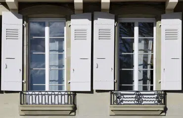 Zwei klassische französische Fenster mit weißen Fensterläden und filigranen schmiedeeisernen Balkongittern in einer Steinfassade.