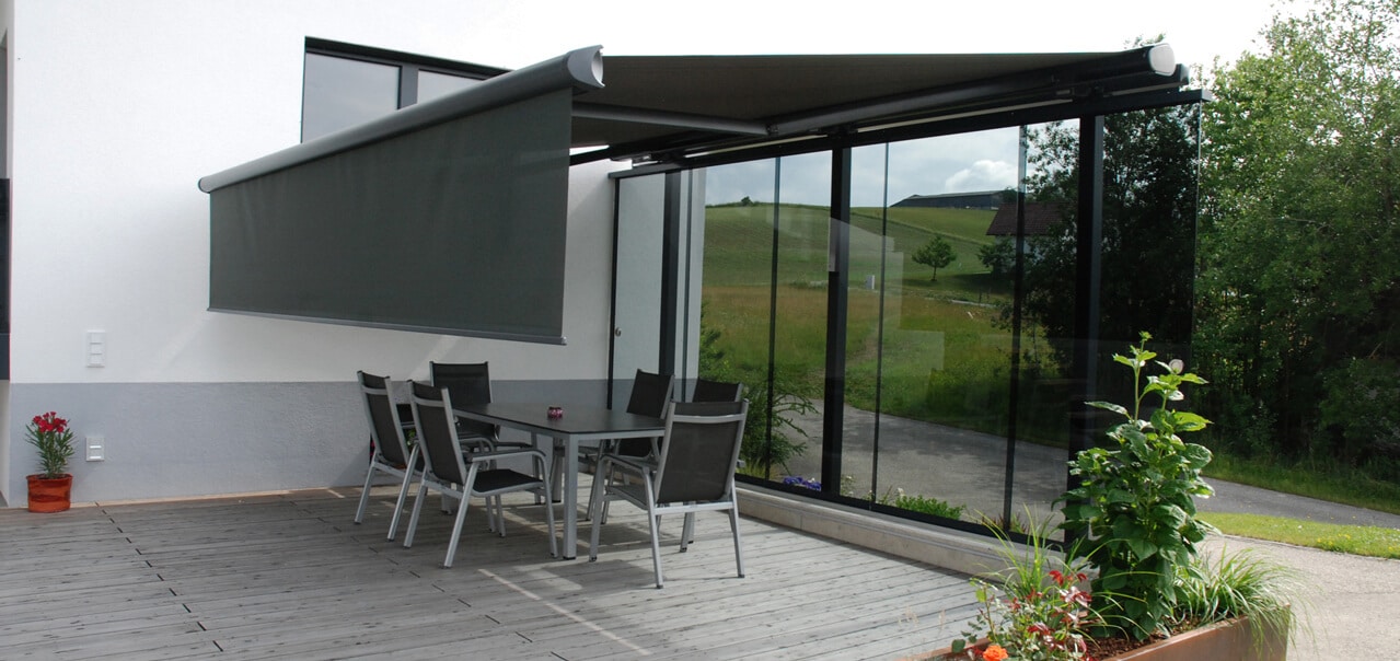 Terrasse mit moderner Glaswand als Windschutz. Eine graue Markise mit seitlichem Rollo spendet Schatten. Davor steht ein Tisch mit vier Stühlen, im Hintergrund grünes Hügelland.