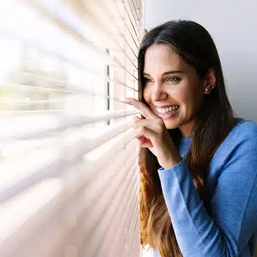 Fröhliche junge Frau blickt durch Jalousien eines neuen waku Fensters.