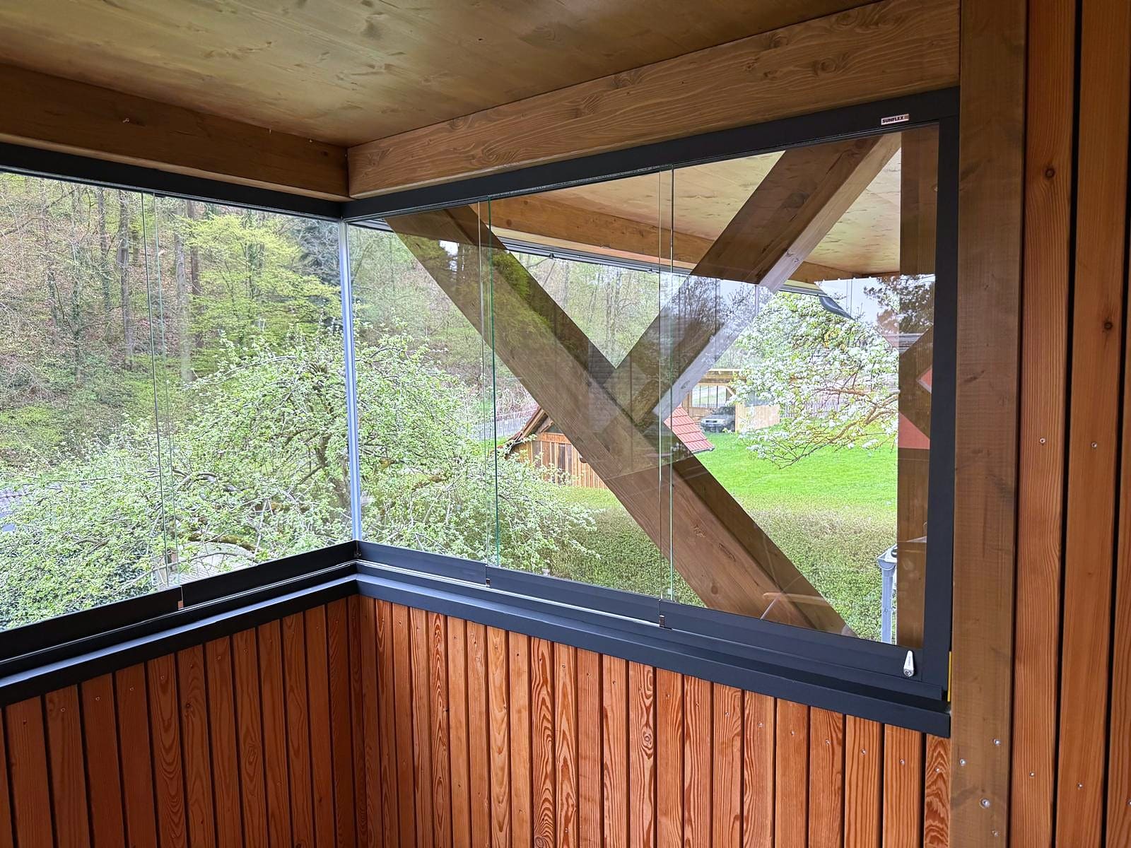 Rahmenlose Ganzglas-Schiebefenster über Eck auf der Lärchenbrüstung eines Holzbalkons, Laufschienen in Anthrazit, Blick in den Garten, Oberösterreich