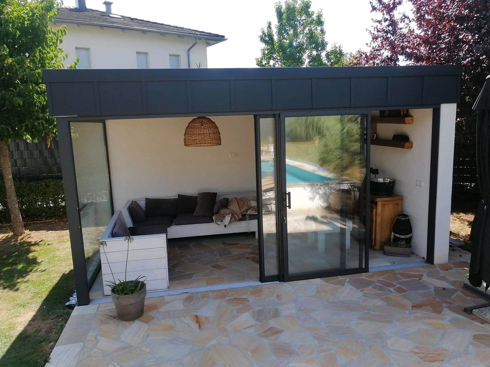 Gartenhaus mit Flachdach, geöffnete Alu-Schiebetüren in DB 703 Dunkelgrau, Lounge innen, Pool spiegelt sich im Glas, Oberösterreich