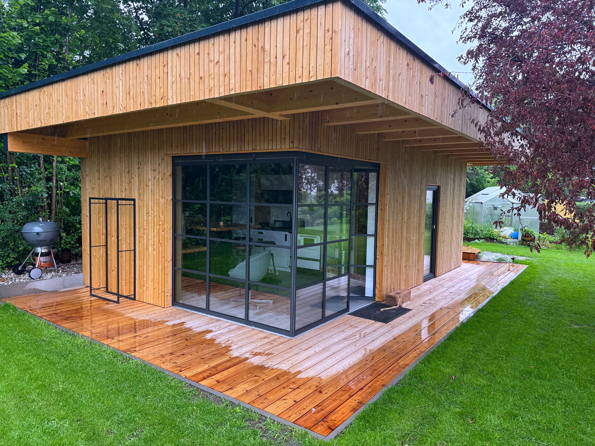 Fertiges Gartenhaus mit Lärchenschalung, Flachdach und Holzterrasse, Schiebe-Dreh-Verglasung mit dunkelgrauen Sprossenrahmen über Eck, Oberösterreich