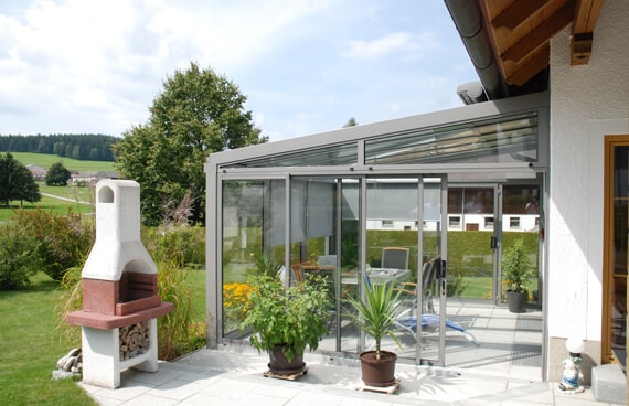 Das Bild zeigt einen Sommergarten mit Glasfronten, Pflanzen und Sitzmöbeln, eingebettet in einen gepflegten Garten.