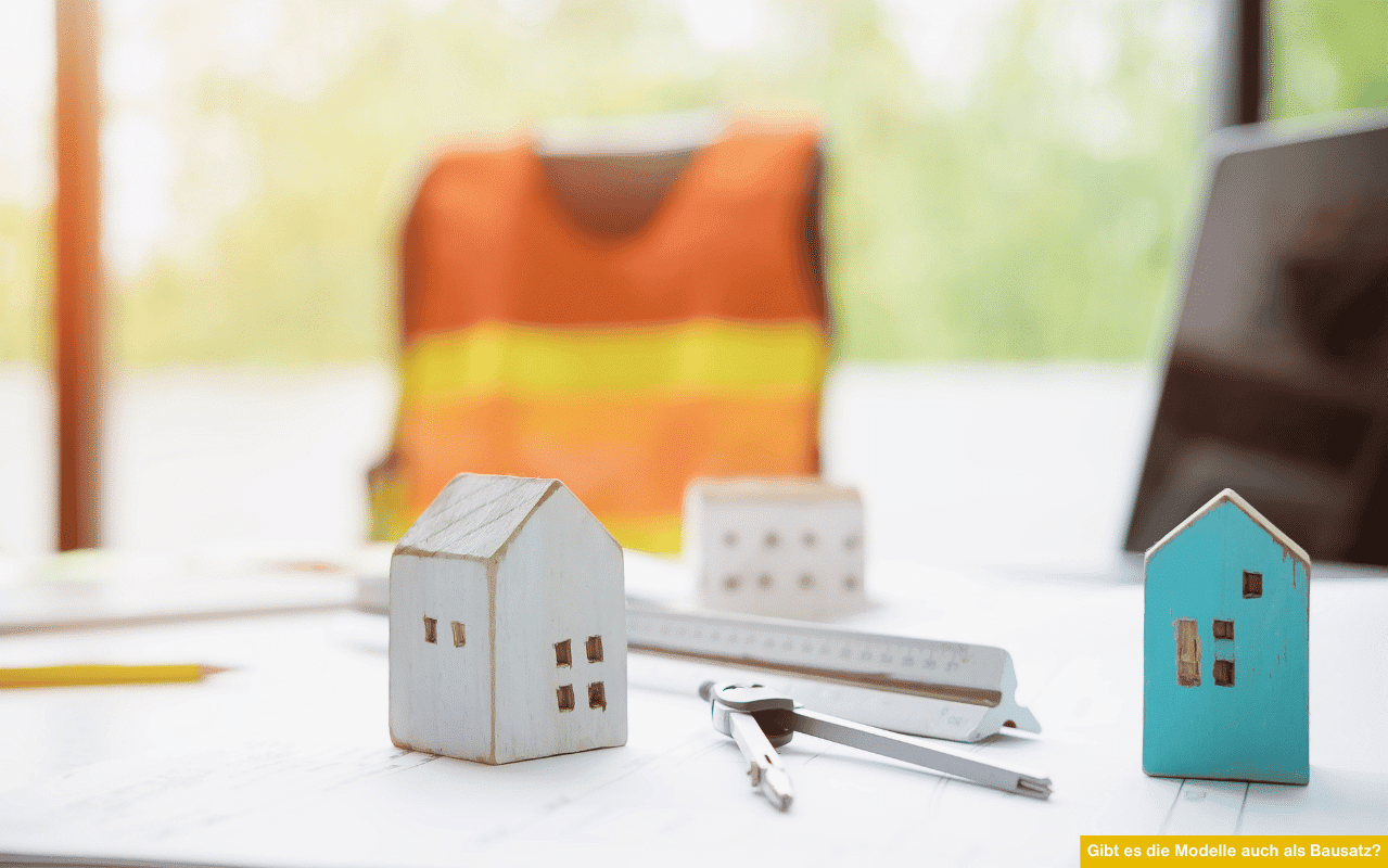 Das Bild zeigt zwei kleine Holzhausmodelle, ein Lineal und einen Zirkel auf einem Tisch mit Bauplänen. Im Hintergrund liegt eine orangefarbene Sicherheitsweste - das Motiv symbolisiert Planung und Maßanfertigung statt Bausatz.