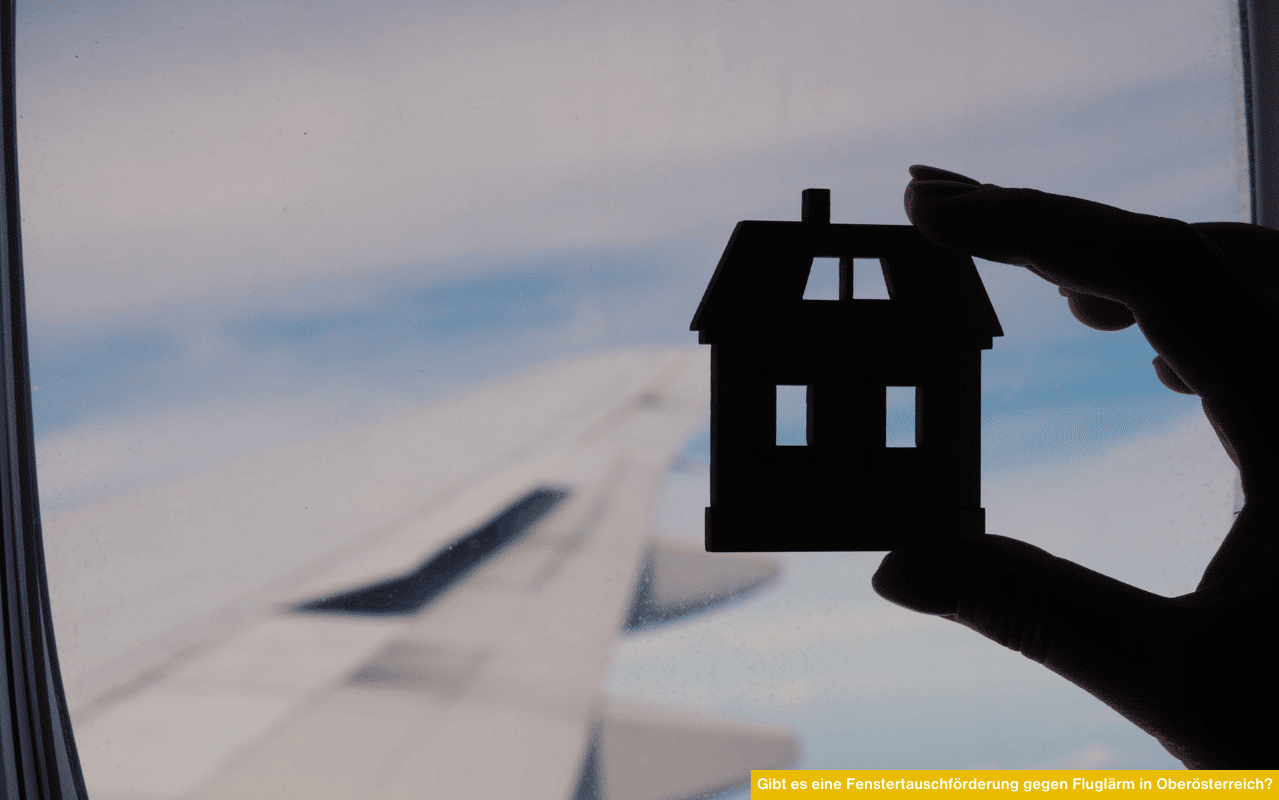 Ein Miniaturhaus wird vor einem Flugzeugfenster hochgehalten, im Hintergrund ist der Himmel mit Flugzeugflügel sichtbar.