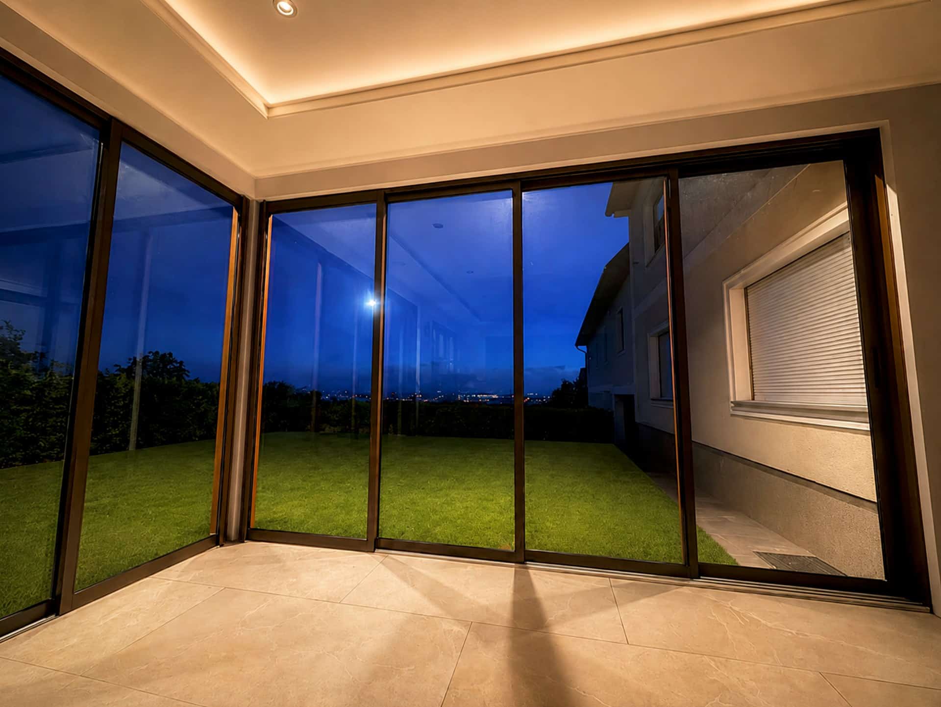 Innenansicht einer Terrassenverglasung mit Glasschiebeelementen in Holzoptik am Abend, Blick über Eck in den Garten, indirekte Deckenbeleuchtung