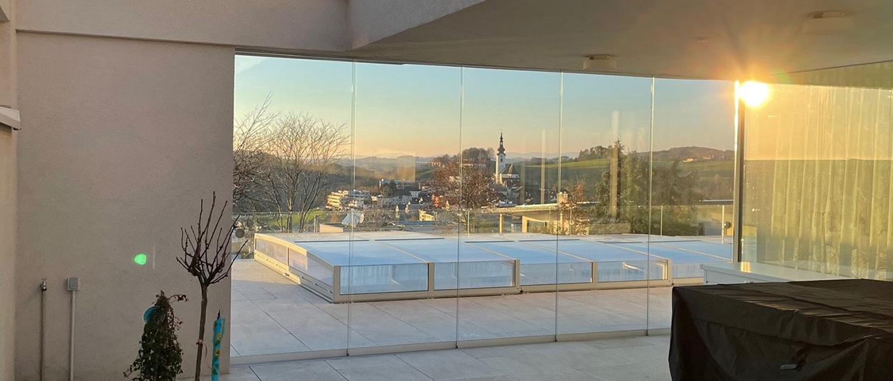Fünfteilige, nahezu rahmenlose Glasschiebewand auf einer überdachten Terrasse. Die transparenten Glaselemente schützen vor Wind und ermöglichen einen freien Blick auf die umliegende Landschaft bei tief stehender Sonne.