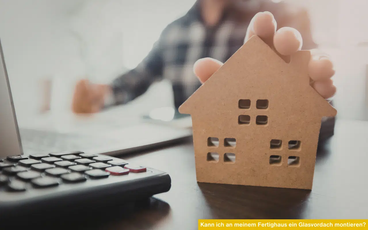 Ein hölzernes Hausmodell steht neben einem Taschenrechner auf einem Schreibtisch als Symbol für die Planung am Fertighaus.
