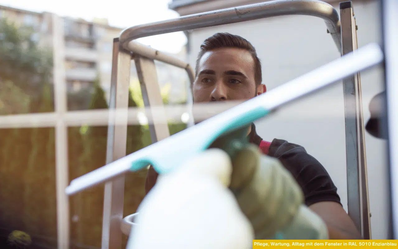 Ein Fachmann säubert ein Fenster in RAL 5010 Enzianblau. Die Szene verdeutlicht, wie regelmäßige Pflege und Wartung der Rahmen, Dichtungen und Beschläge die Lebensdauer und das Aussehen der Fenster verbessern.