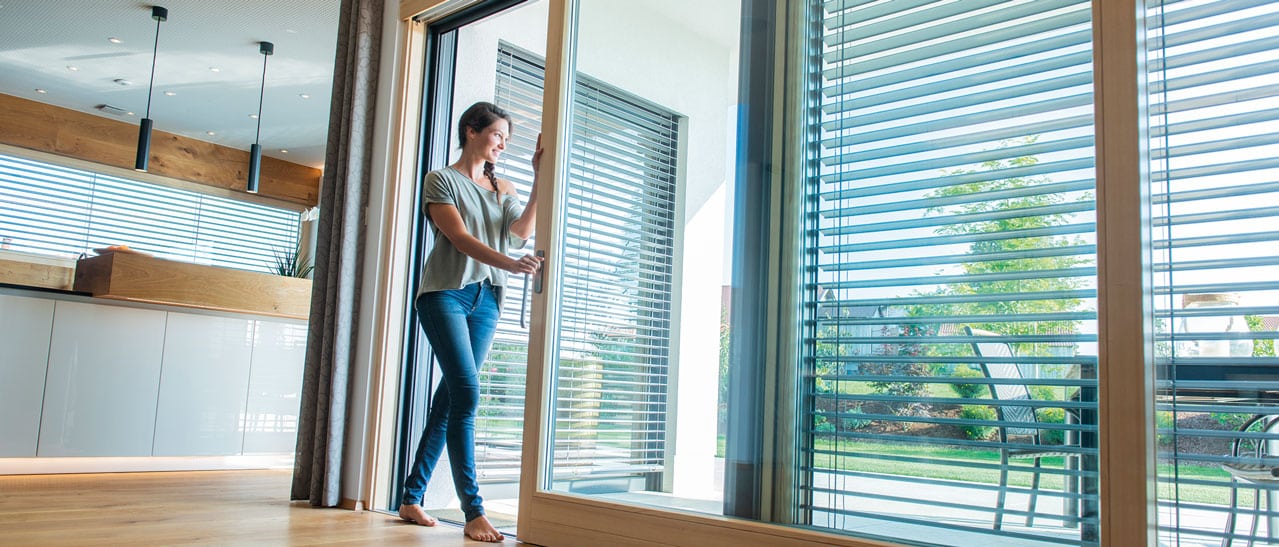 Frau öffnet Schiebeflügel einer modernen Glasfront und genießt den Zugang zur sonnigen Terrasse mit Blick ins Grüne.