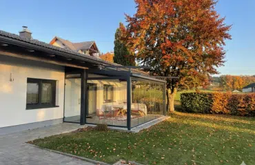 Eine Außenansicht eines gläsernen Wintergartens an einem weißen Haus, umgeben von einem herbstlichen Garten mit einem prachtvollen, bunt gefärbten Baum.