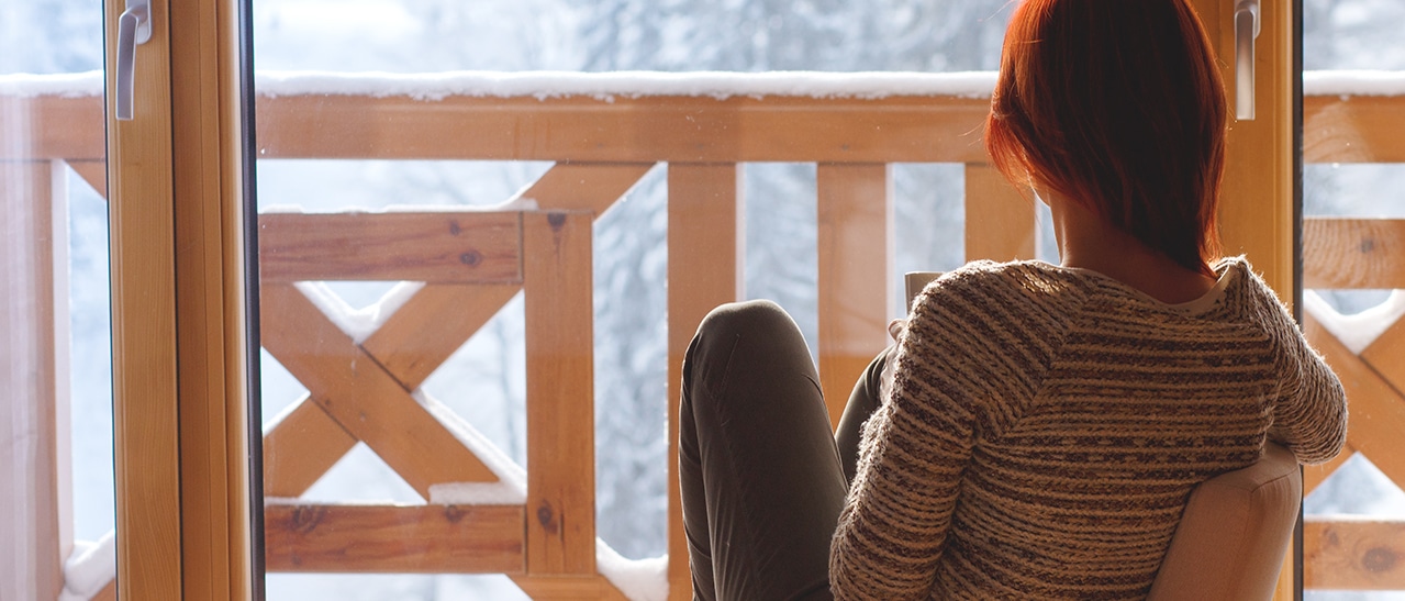 Die Aufnahme zeigt Holz-Alu-Balkontüren, durch die man auf einen Balkon mit Schneelandschaft blickt. Eine Frau sitzt entspannt im warmen Raum und genießt die winterliche Aussicht.