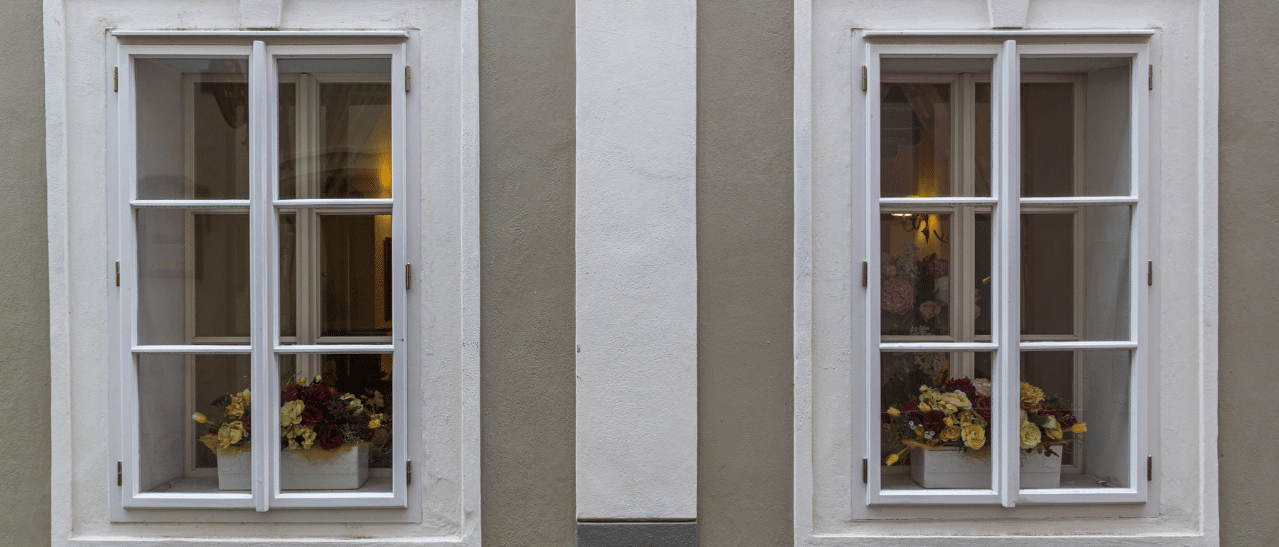 Zwei symmetrisch angeordnete Holzfenster mit Sprossen und Blumen am Fensterbrett - klassischer Fassadenstil mit charmanten Details.