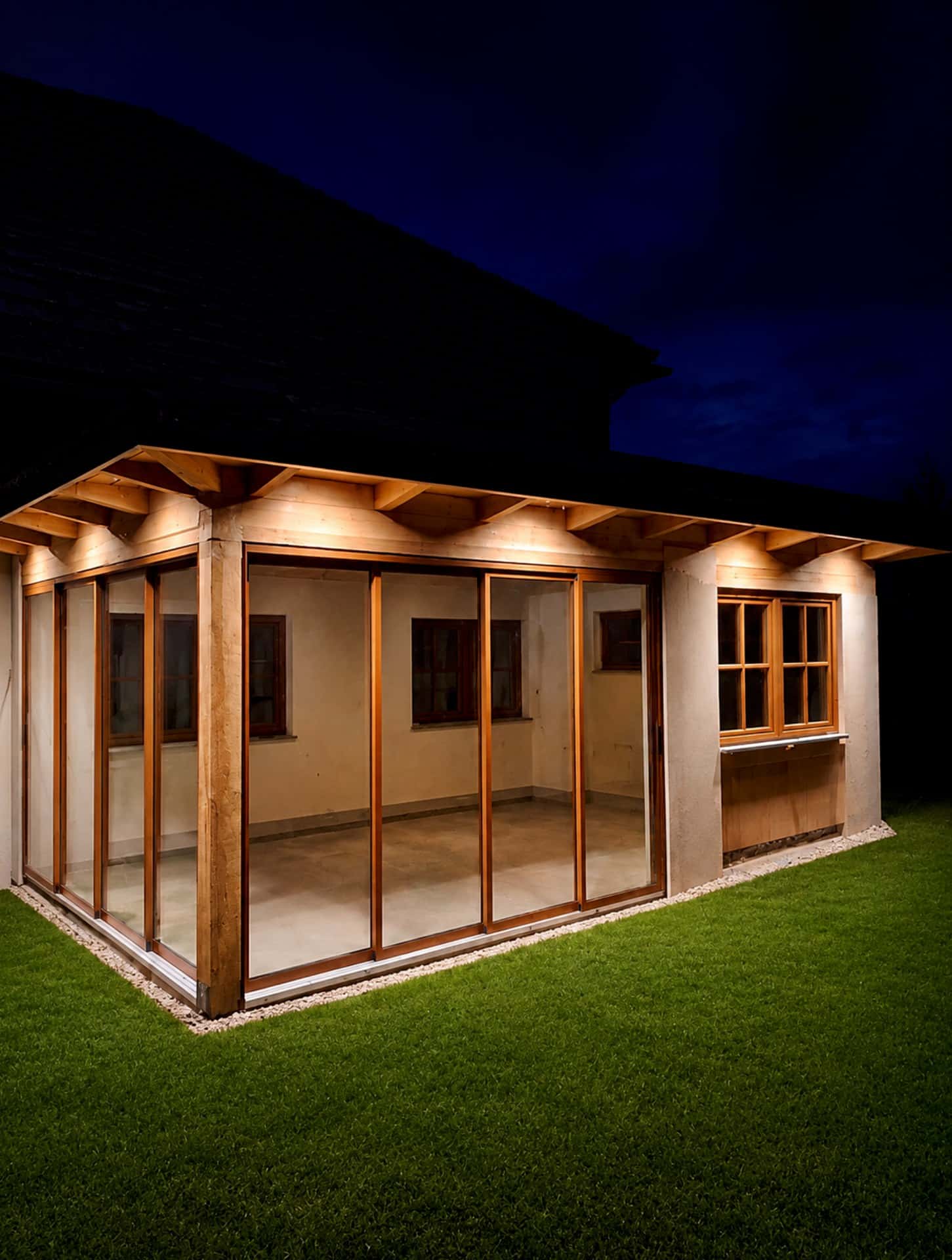 Beleuchtete Holzkonstruktion einer Terrassenverglasung mit Glasschiebeelementen in Holzoptik am Abend, Außenansicht über Eck, Oberösterreich