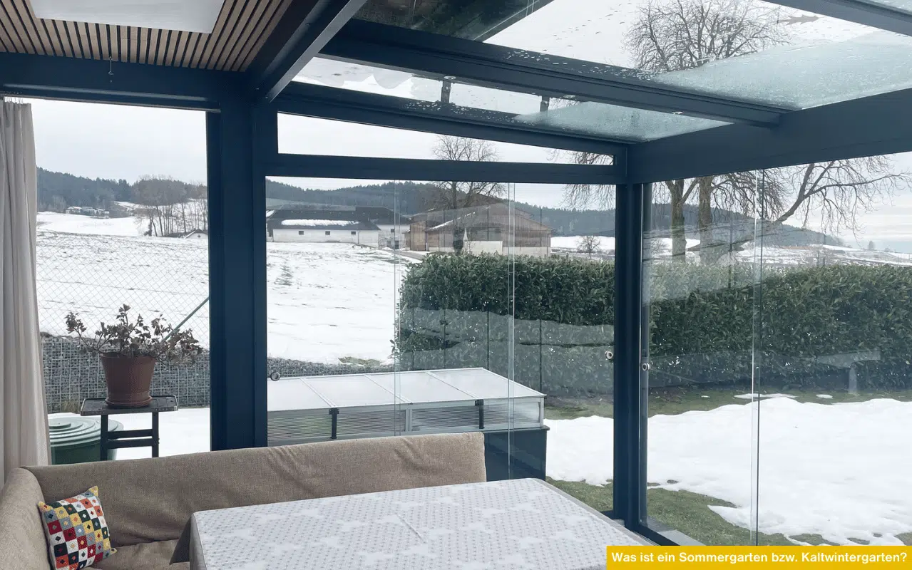 Innenansicht eines Kaltwintergartens mit dunklem Alurahmen und großen Glasscheiben. Eine gemütliche Sitzecke ist im Vordergrund, durch die Verglasung sieht man eine winterliche Landschaft mit Schneeflächen und Hecke.