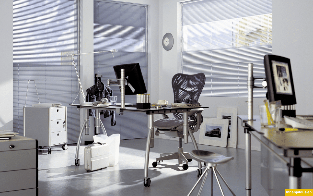 Das Bild zeigt ein modernes Büro mit großen Fenstern, die durch silberne Innenjalousien verschattet werden. Die Jalousien regulieren das Tageslicht und sorgen für eine angenehme Arbeitsatmosphäre.
