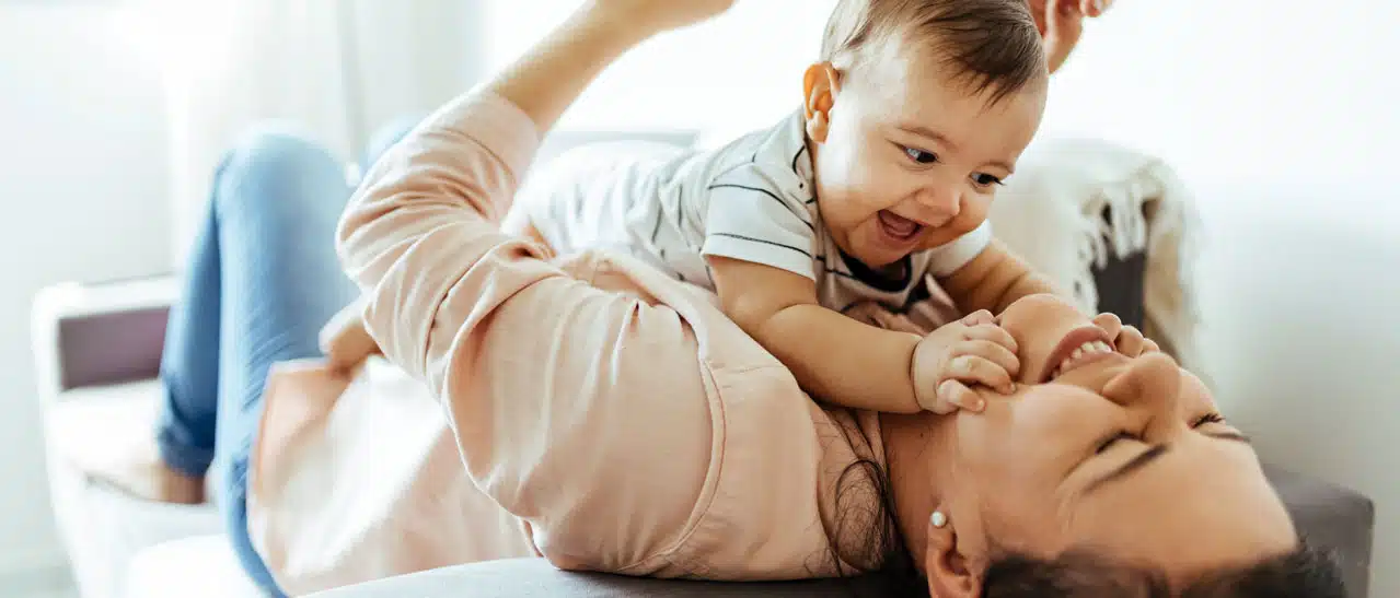 Junge Frau mit Baby auf der Couch, im Hintergrund die Fenster, wo sie die Gläser ausgetauscht hat und jetzt wertvolle Heizkosten spart. 