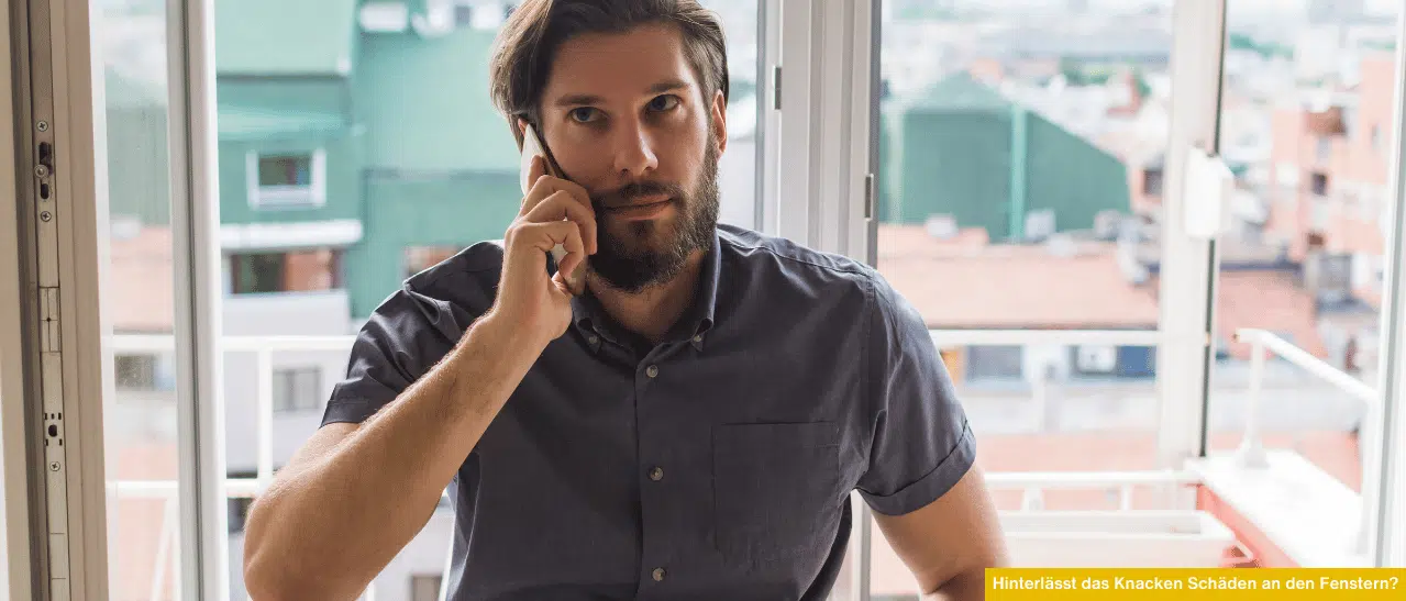 Junger Mann sitzt ratlos vor seinem geöffneten Fenster mit Mobiltelefon in der Hand, kontaktiert Fensterfirma wegen Fenster knacken.