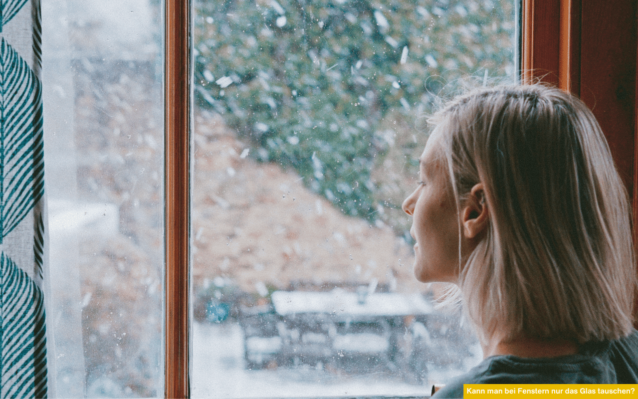 Kann man bei Fenstern nur das Glas tauschen zeigt Frau, die nachdenklich bei Schneefall durch ein Fenster mit Holzrahmen blickt.