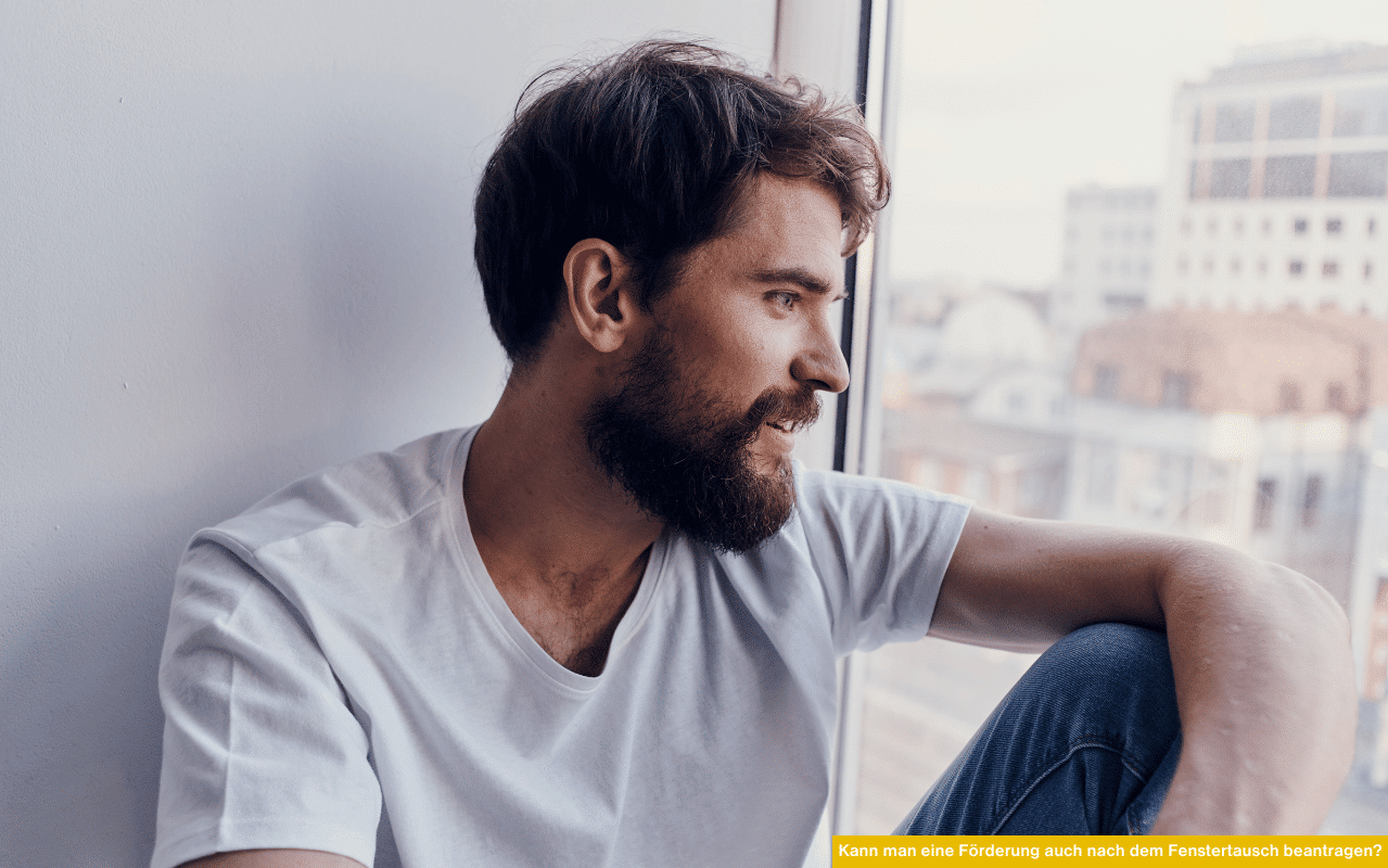 Ein junger Mann mit Bart sitzt in einem hellen Raum am Fenster, schaut nach draußen und wirkt nachdenklich.